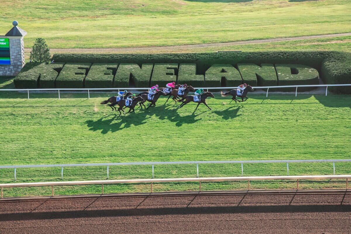The Best of Lexington, KY: Iconic Stops + Hidden Gems - 3 A group of jockeys riding horses races on a grass track at Keeneland in Lexington, the Horse Capital, with large trimmed bushes spelling “KEENELAND” in the background.