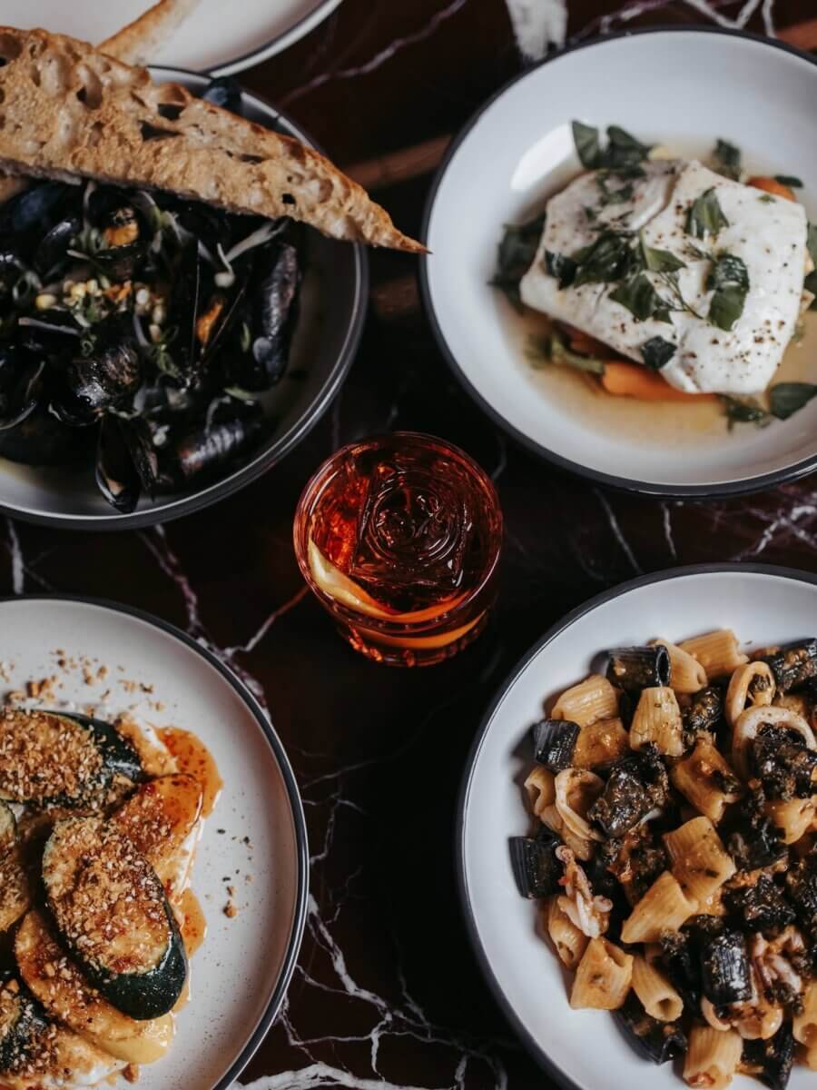 The Best of Lexington, KY: Iconic Stops + Hidden Gems - 8 Four plates of assorted Italian dishes and a red cocktail with a citrus twist are arranged on a dark marble table, capturing the essence of discovering Lexington’s vibrant dining scene in the Horse Capital of the World.