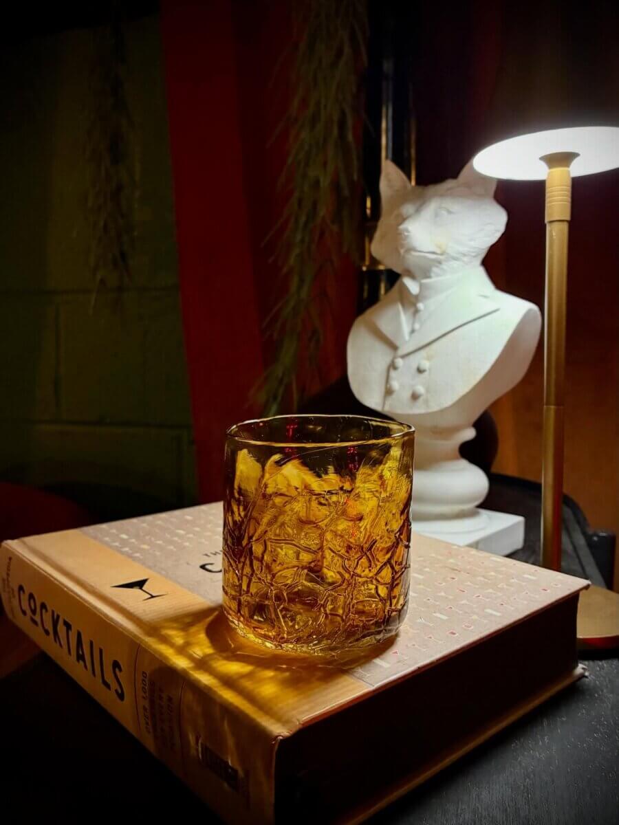 The Best of Lexington, KY: Iconic Stops + Hidden Gems - 9 A glass of amber liquid sits on a cocktail book next to a white fox statue and a lit desk lamp, evoking the refined charm of Lexington.