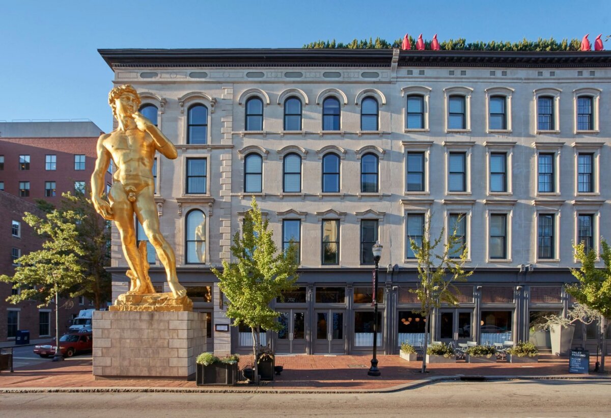 Your Guide to Boutique Hotels in Louisville - 2 A golden statue of David stands in front of a historic four-story building with arched windows and storefronts at street level on a sunny day, capturing the vibrant charm often found near Louisville’s boutique hotels.