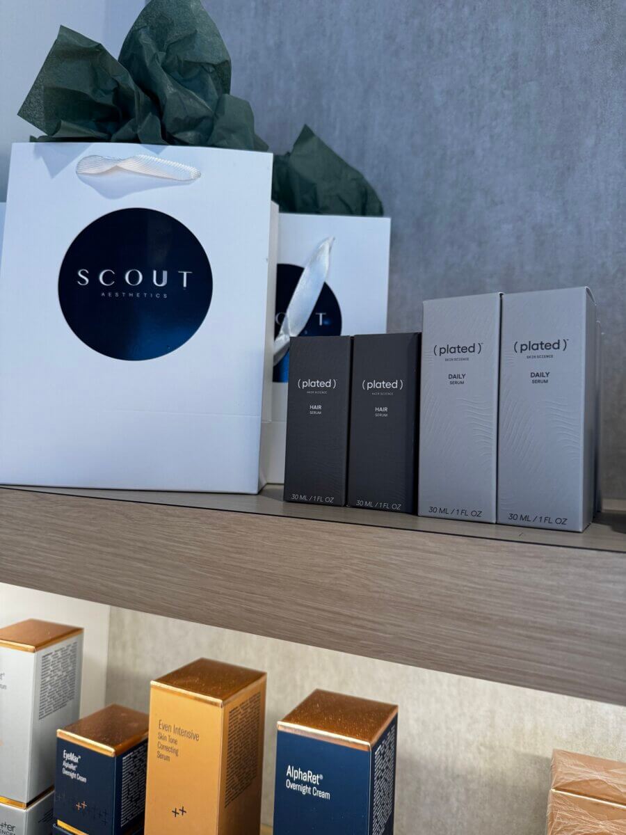 White shopping bags labeled “Scout Aesthetics” and boxed skincare products are displayed on a wooden shelf against a gray wall.