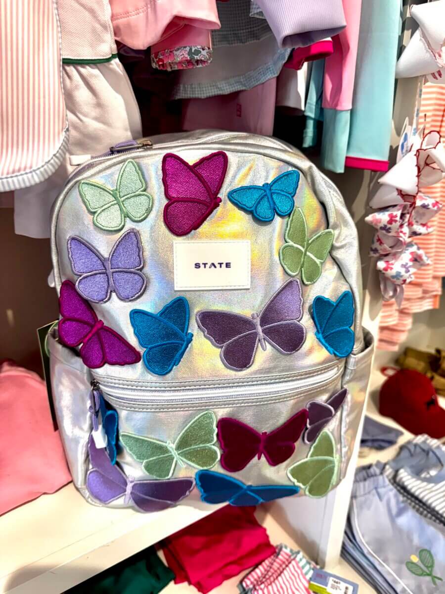 A silver backpack with colorful butterfly appliqués is displayed among children’s clothing in a store.