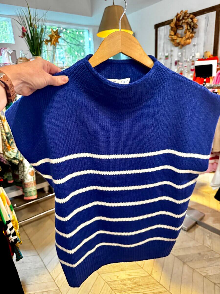 A sleeveless blue knit top with white horizontal stripes is held on a wooden hanger in a clothing store, making it a perfect Fall Preview piece for your wardrobe.