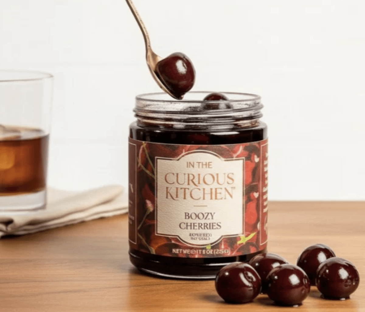 A jar of The Curious Kitchen Boozy Cherries sits on a wooden surface with a spoon lifting a cherry and several cherries and a drink nearby.
