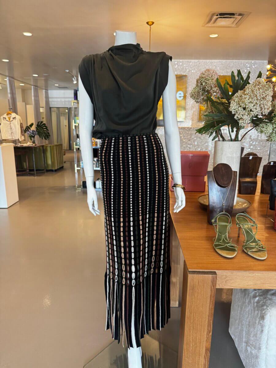 A mannequin displays a black sleeveless top and a long black skirt with vertical white patterns and fringe at the hem, capturing this season’s Fall Preview in a modern boutique setting.