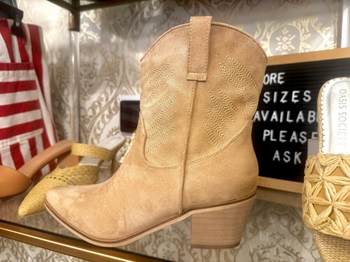 Tan suede cowboy boot with a low wooden heel displayed on a shelf, surrounded by other shoes and a sign stating, “More sizes available, please ask”—a true Louisville FIND.