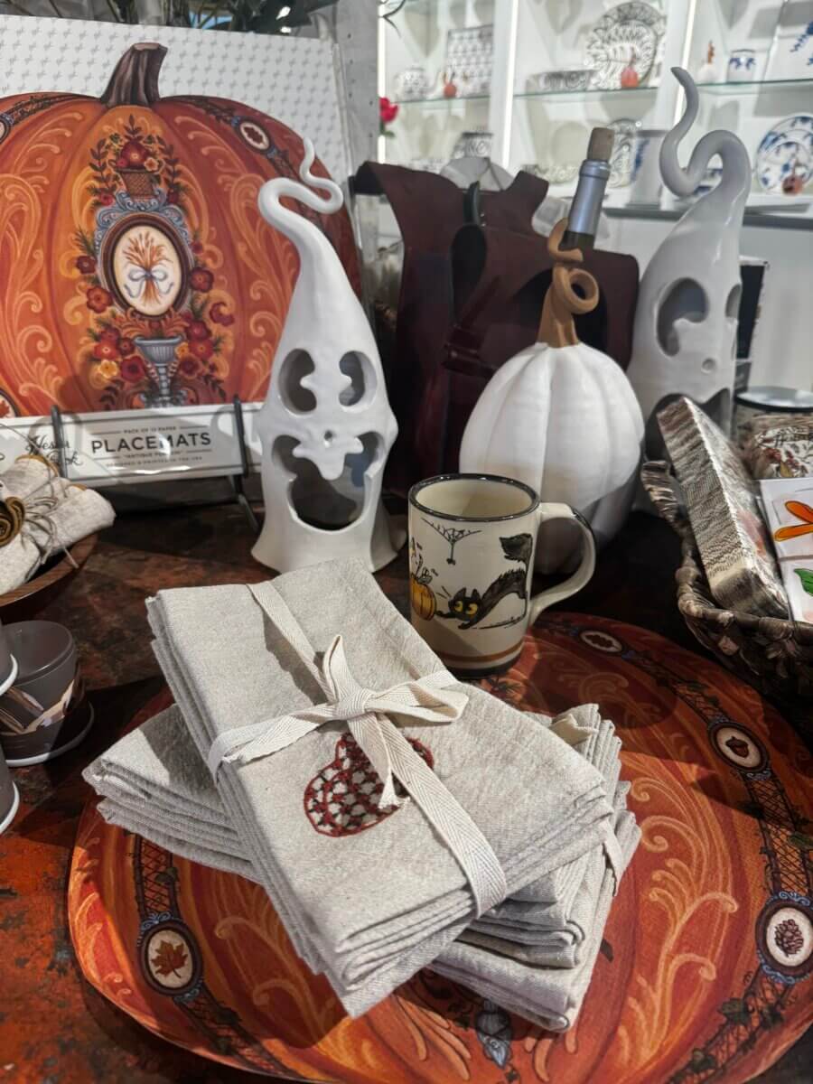 A display of Halloween-themed decor, including white ghost figurines, a pumpkin placemat, folded napkins, a mug, and various tableware items on a wooden surface.