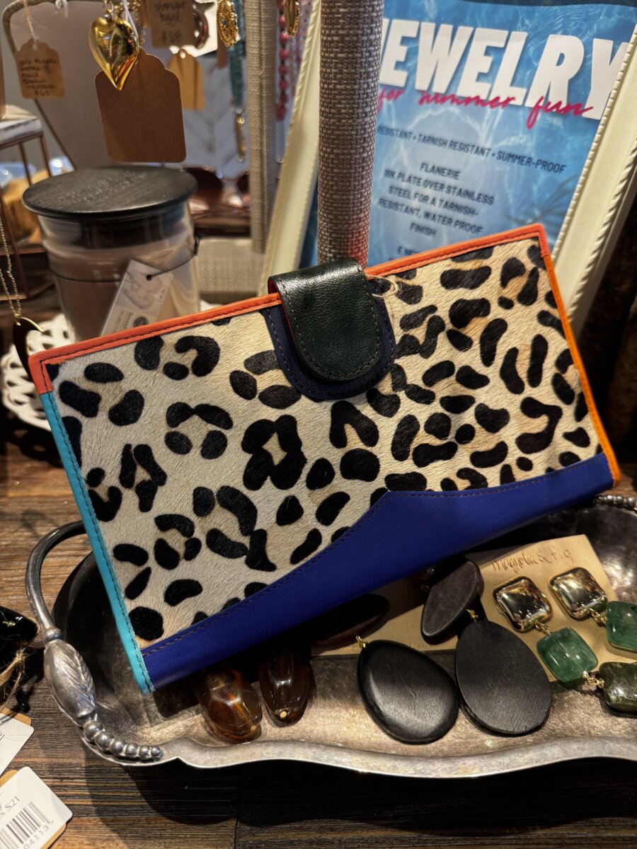 A leopard-print and blue clutch wallet displayed on a tray with polished stones and black jewelry, with a jewelry sign and various items in the background.