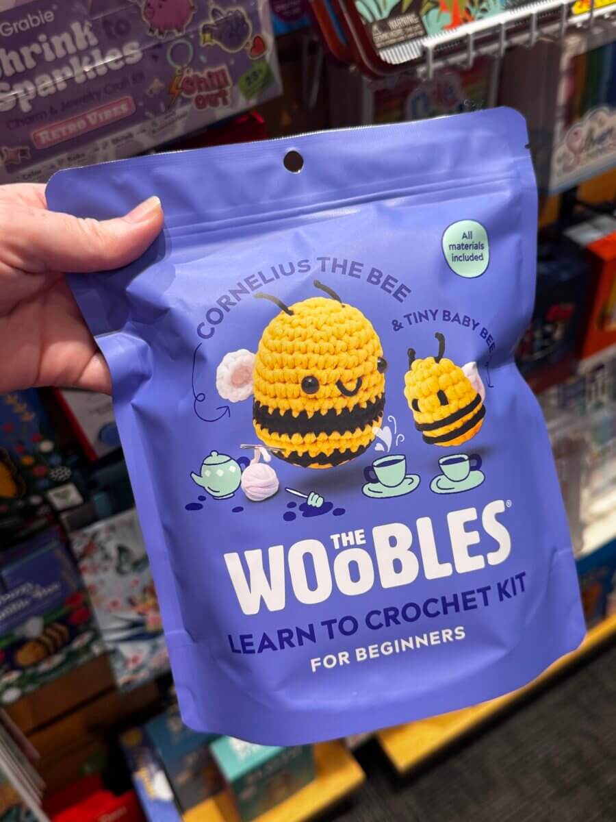 A hand holds a purple bag labeled “The Woobles Learn to Crochet Kit for Beginners,” featuring crocheted bees and tea cups on the packaging.