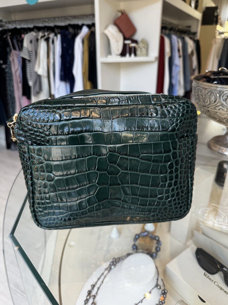Dark green crocodile-embossed leather handbag displayed on a glass shelf in a boutique, with clothing and accessories in the background.