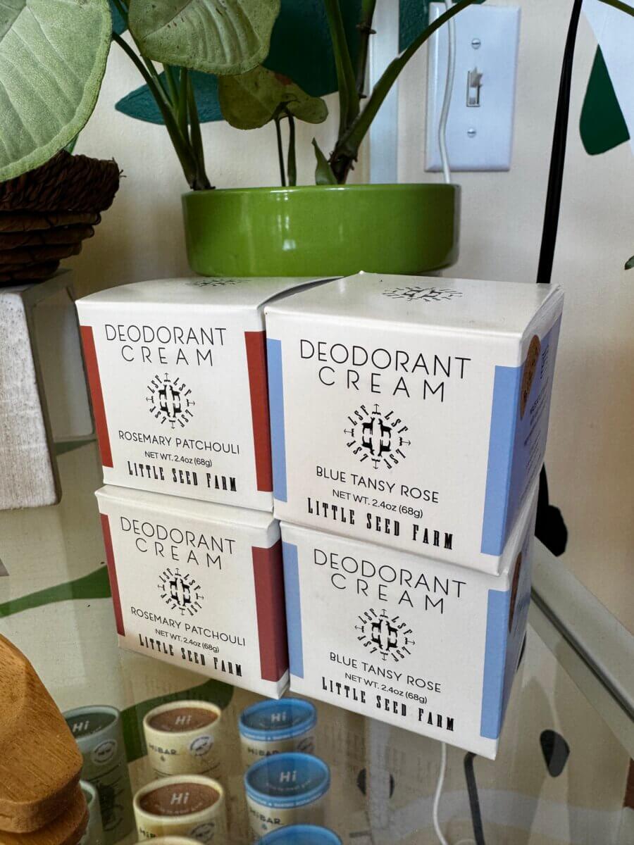 Four boxes of Little Seed Farm deodorant cream—two labeled “Rosemary Patchouli” and two “Blue Tansy Rose”—are neatly stacked on a glass shelf near a potted green plant and a light switch, making this display one of our top Home Must Haves from the Louisville FINDS Fall Preview.
