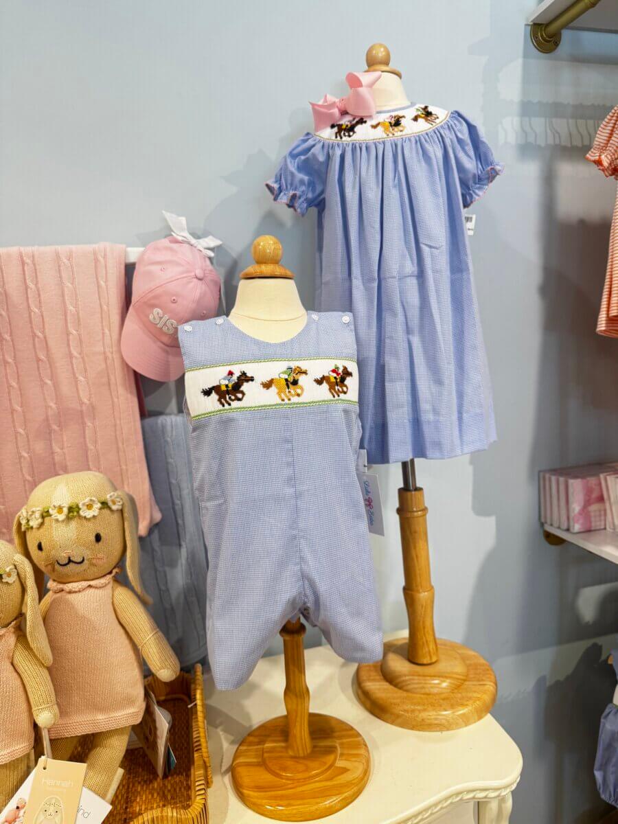 Two child-sized mannequins display matching blue embroidered outfits; one is a romper, the other a dress—a charming duo. A pink cap, pink blanket, and two dolls add sweet touches to this delightful spread of Louisville finds on a white table.