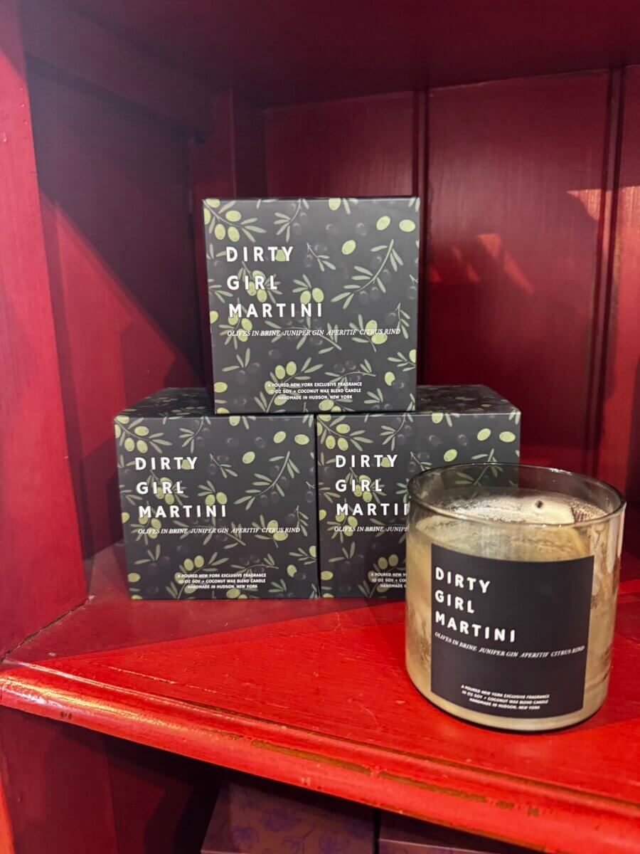 Four “Dirty Girl Martini” scented candles are displayed on a red shelf; three are boxed, and one is unboxed in a glass container.