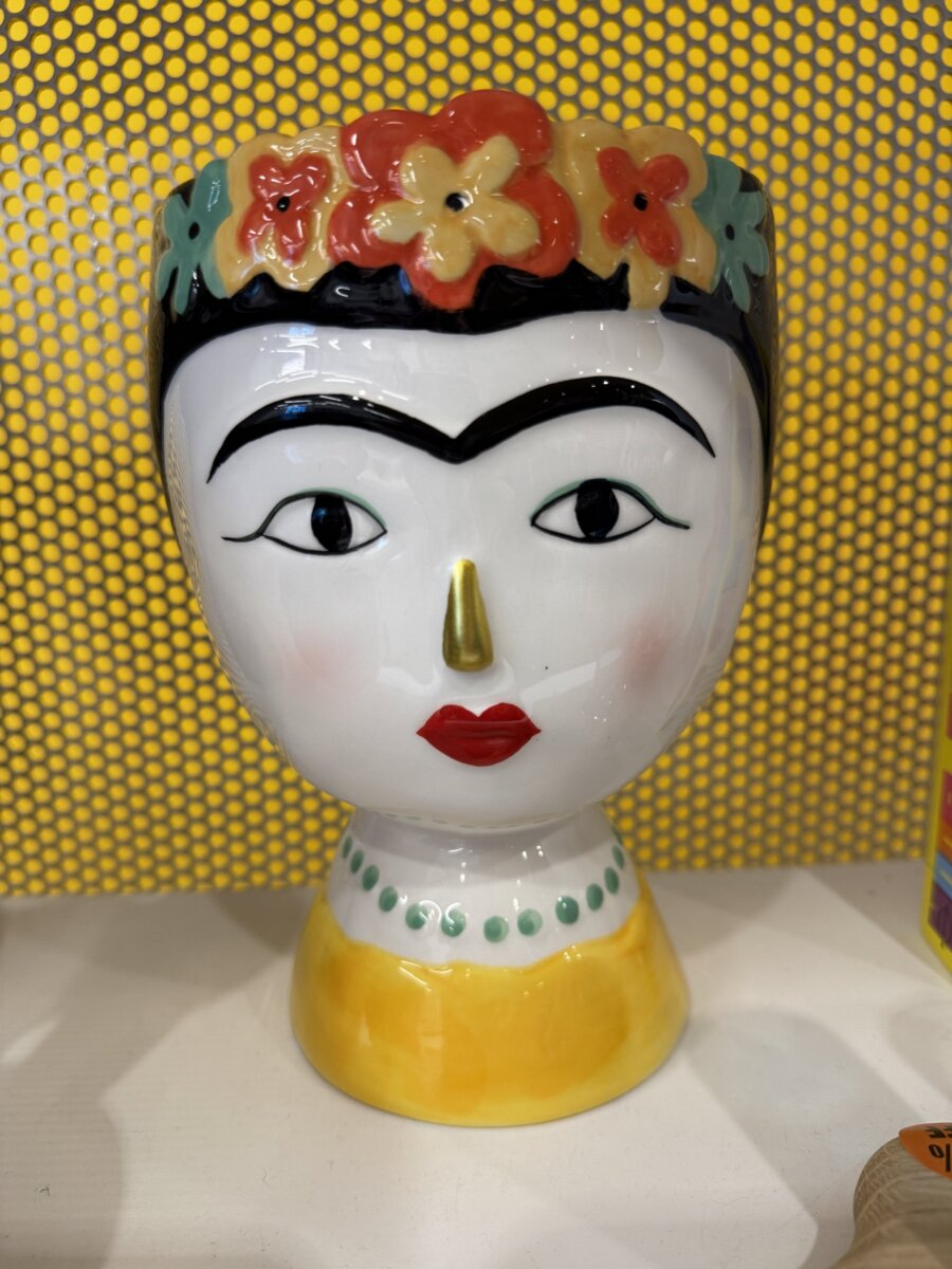 Ceramic head-shaped vase with colorful flowers on top, bold eyebrows, red lips, and a yellow base, displayed against a yellow dotted background.