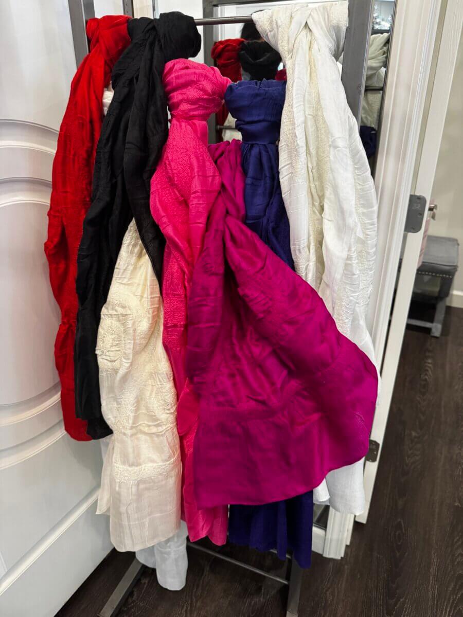Several colorful scarves, including red, black, white, pink, and purple, hang on a metal rack in a room with dark wood floors.