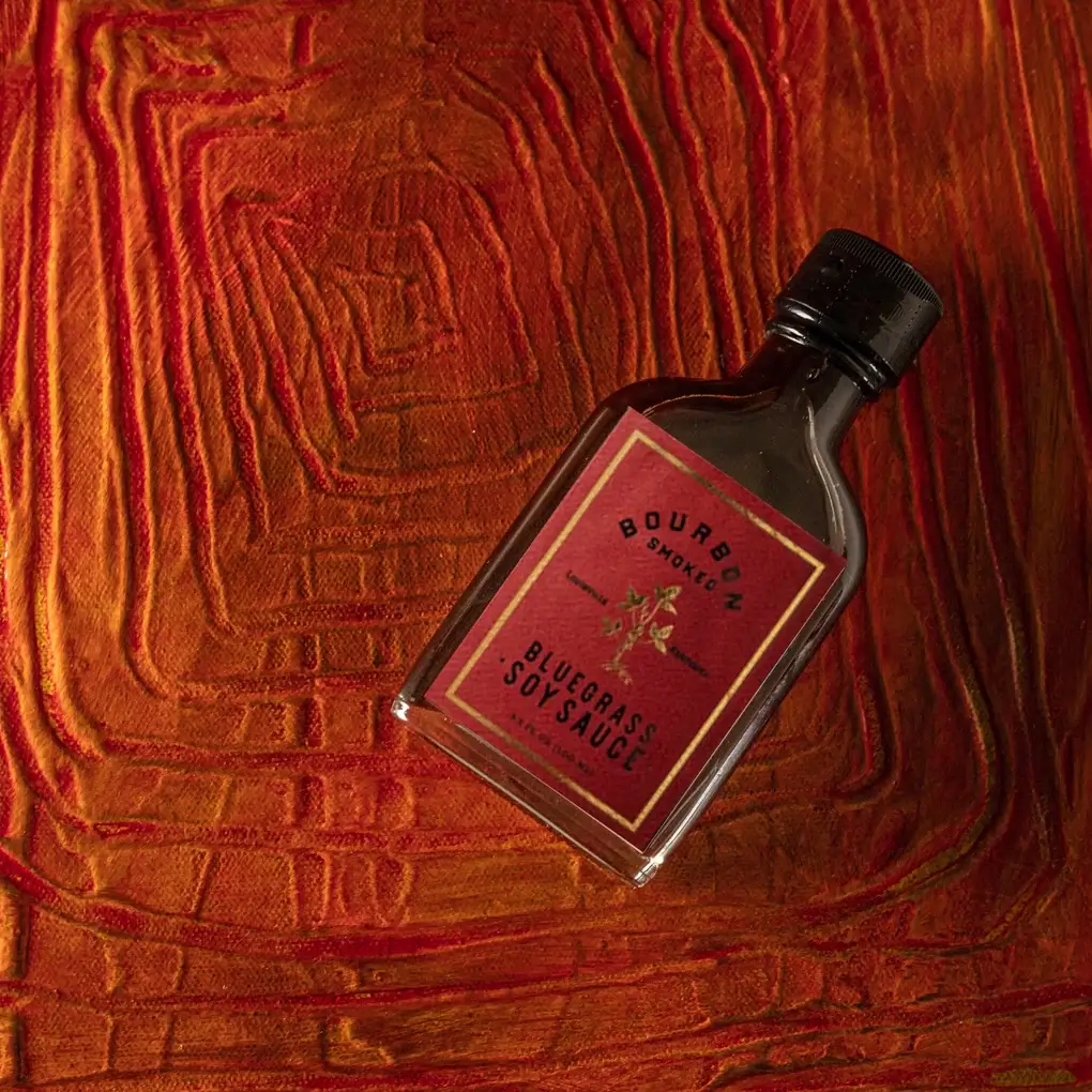 A small glass bottle of Bourbon Smoked Bluegrass Soy Sauce with a red label and black cap, placed on a textured red and orange surface that evokes the warm hues of fall in Louisville.