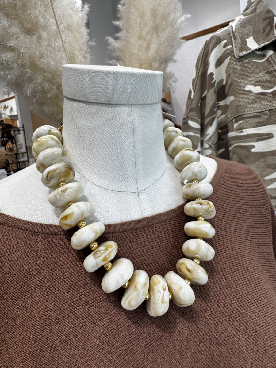 A mannequin wearing a chunky white and gold marbled bead necklace and a brown top; a camouflage jacket and pampas grass are in the background.