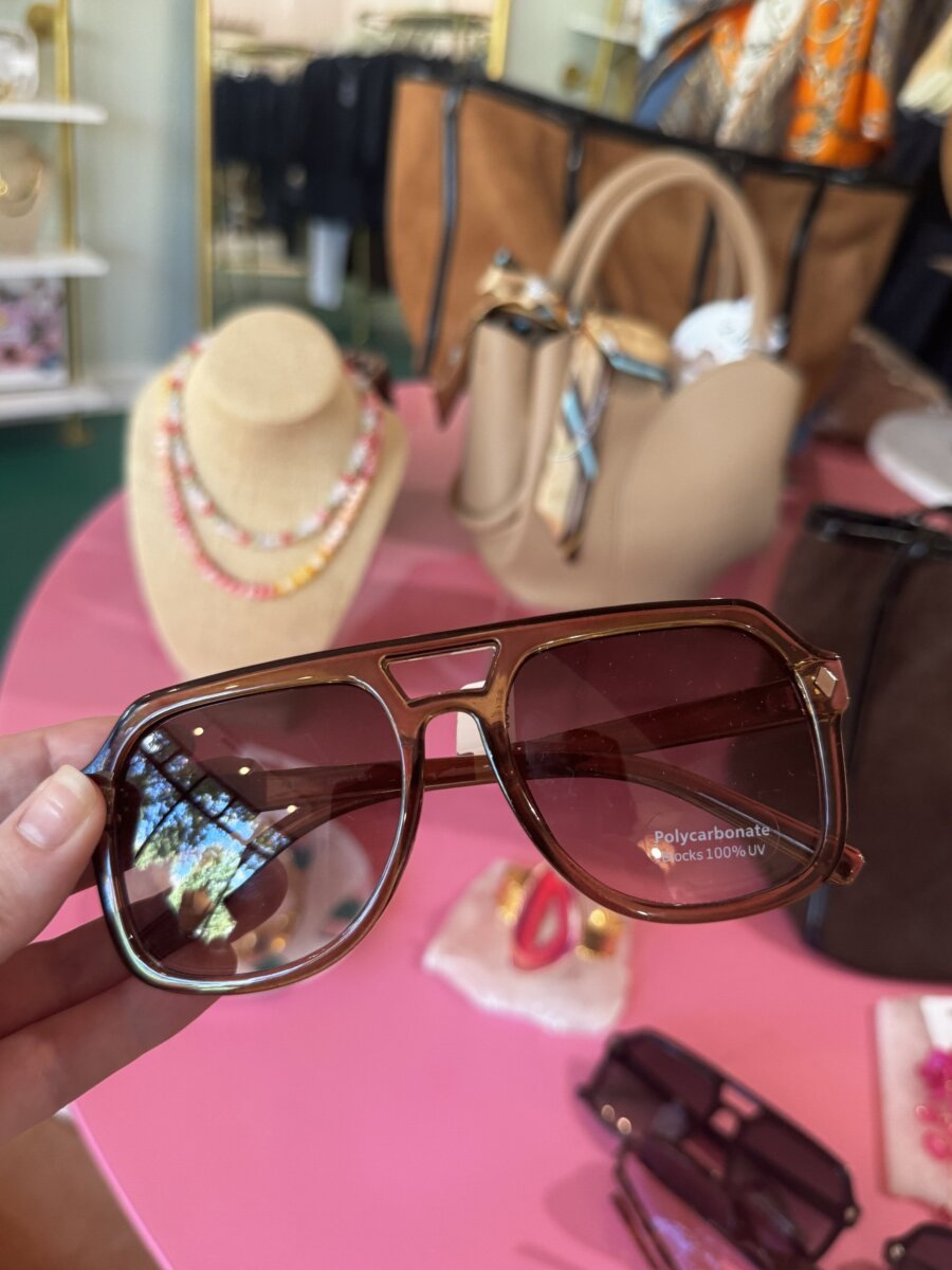 A hand holds brown oversized sunglasses with a polycarbonate label; purses, a beaded necklace, and accessories are displayed on a pink table in the background.