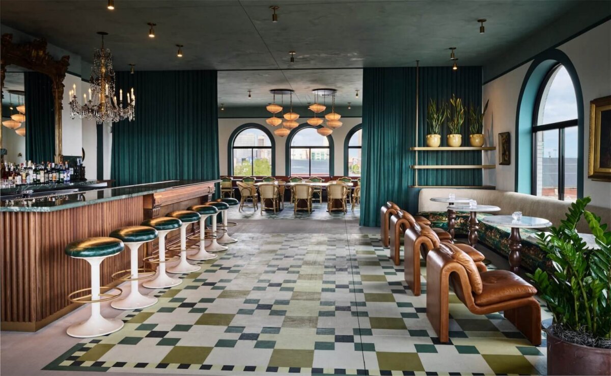 Your Guide to Boutique Hotels in Louisville - 5 A stylish bar in Louisville with green barstools, a chandelier, leather chairs, patterned floor tiles, and arched windows offering city views—a perfect stop for guests staying at nearby boutique hotels. Tables and plants decorate the modern interior.
