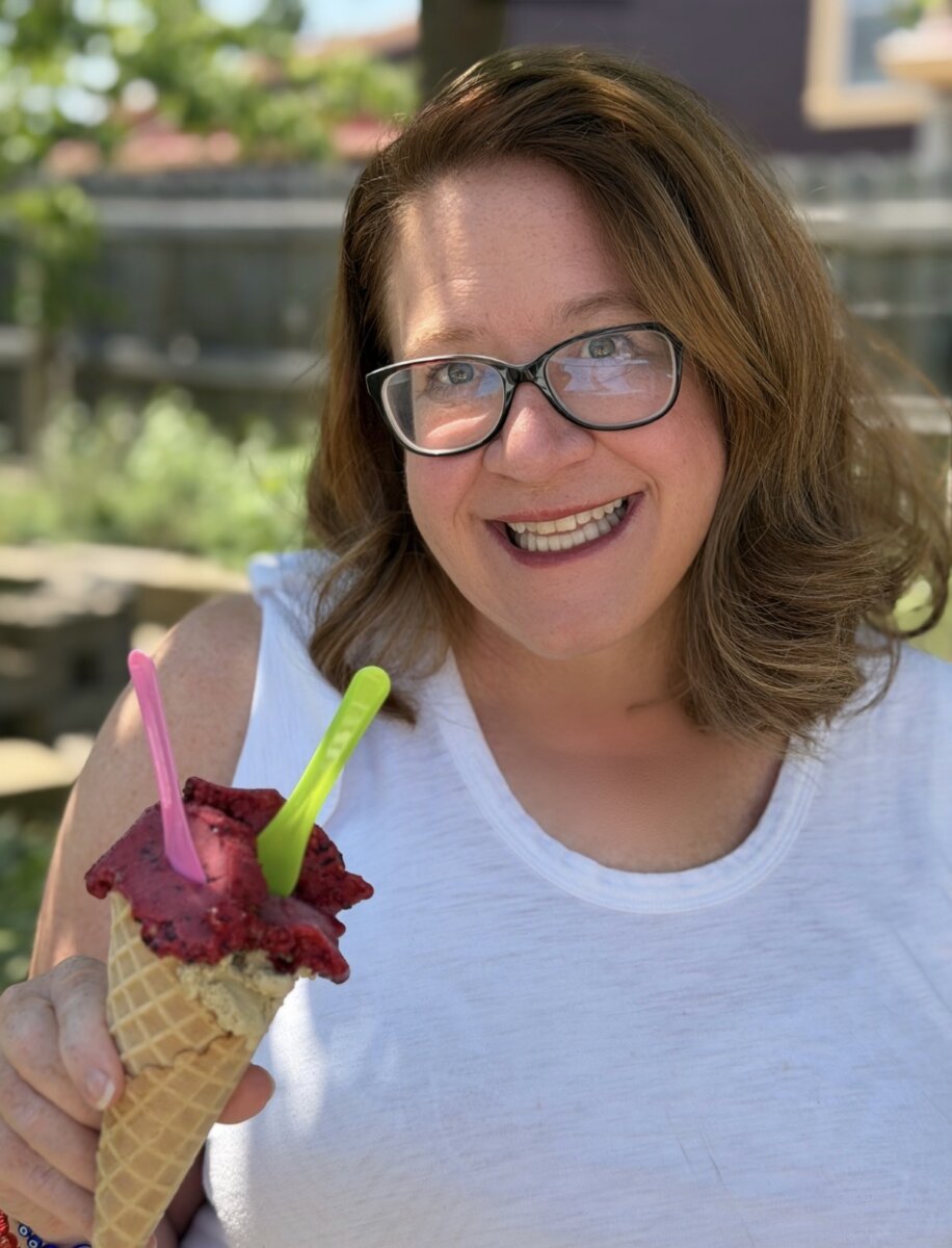 Meet the Art Historian Behind Louisville’s Gelato Gilberto - 2 Woman in glasses and a white shirt smiles outdoors while holding a waffle cone with two scoops of ice cream and two colorful spoons, savoring the simple pleasures of the sweet life.