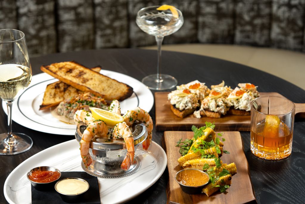3 New Louisville Restaurants to Try (Plus 3 New Menus!) - 5 A table with shrimp cocktail, toasted bread, seafood-topped crostini, salad, three cocktails, and dipping sauces on plates and wooden boards—perfect for sampling the creative offerings found at new restaurants and Louisville restaurants debuting exciting new menus.