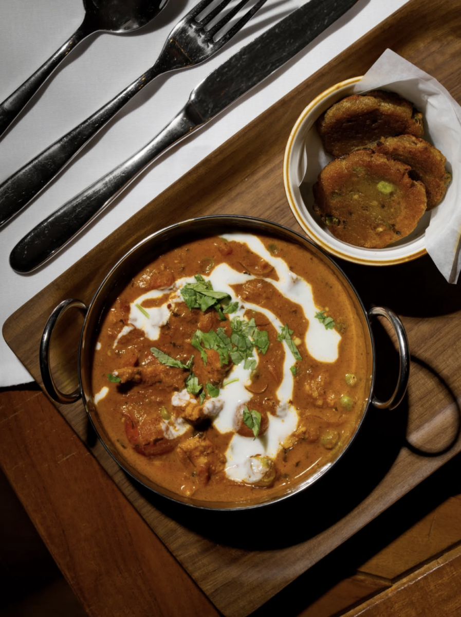 3 New Louisville Restaurants to Try (Plus 3 New Menus!) - 6 A bowl of curry garnished with cream and cilantro is served on a wooden tray with a side of fried patties, accompanied by a fork, knife, and spoon—a tempting dish that highlights the vibrant flavors found at new restaurants in Louisville.