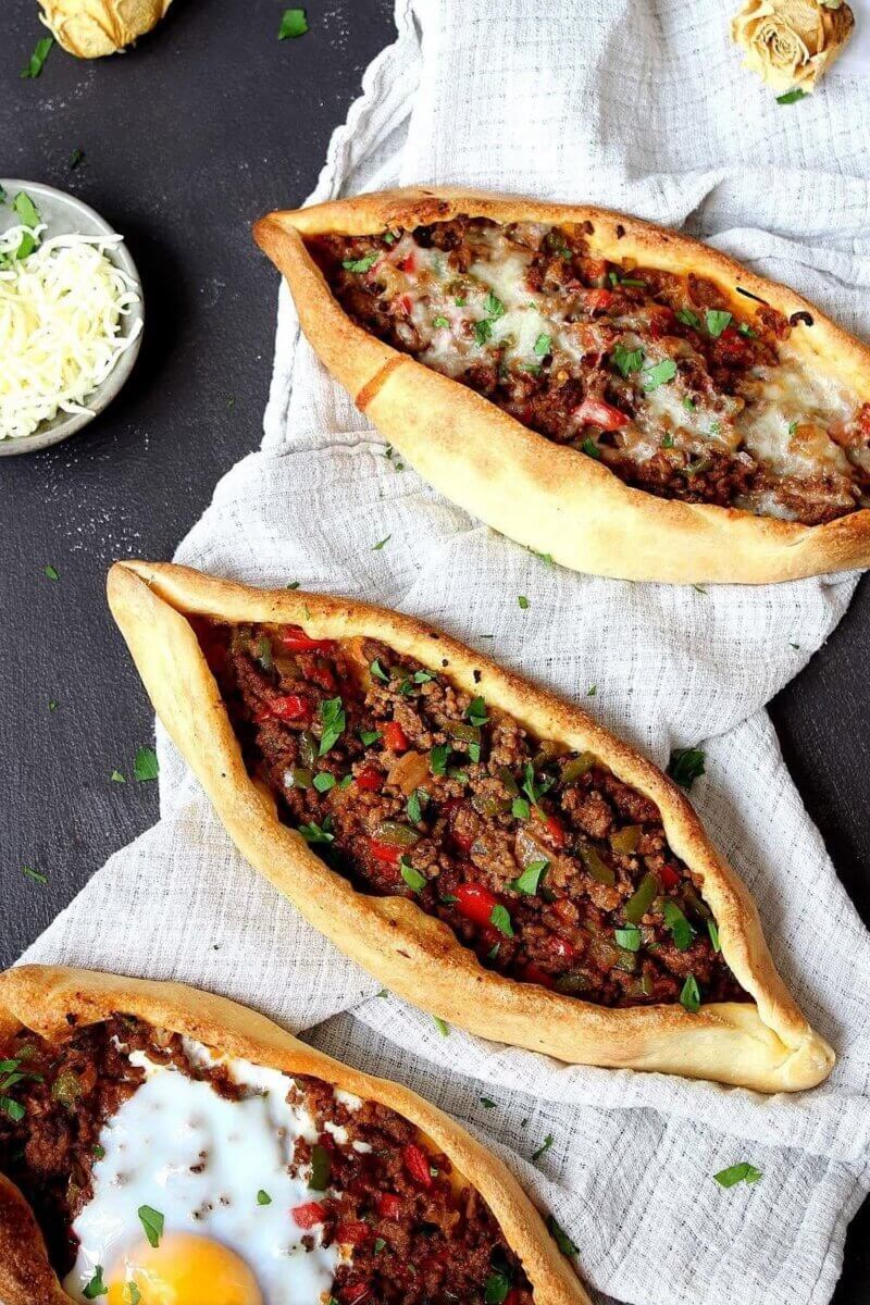 Three boat-shaped Turkish pide breads topped with minced meat, herbs, and vegetables; one has melted cheese and another is crowned with a cooked egg. A bowl of grated cheese sits nearby—a delicious example of what you can expect from the new Louisville restaurants making waves in the city’s dining scene.