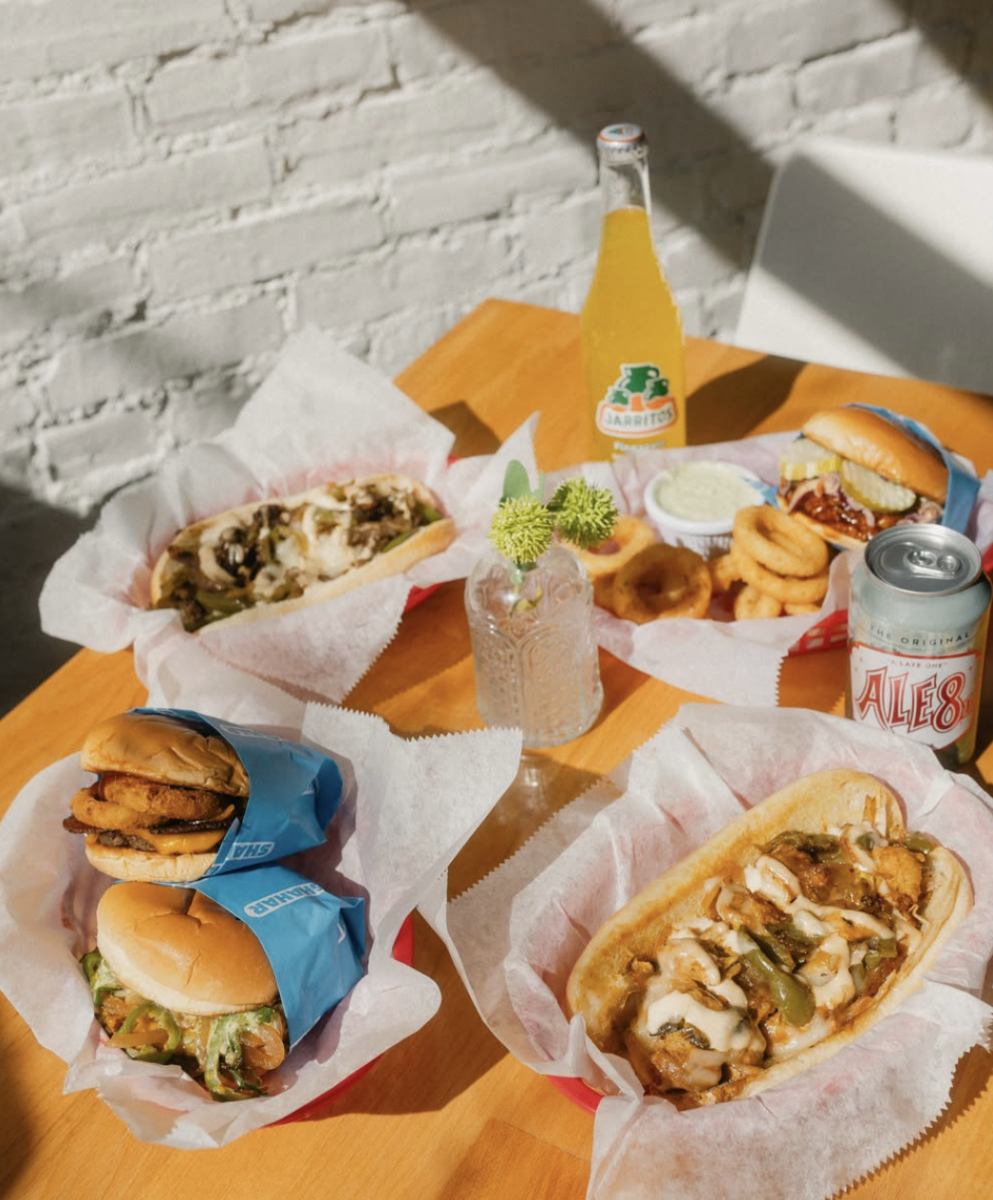 3 New Louisville Restaurants to Try (Plus 3 New Menus!) - 7 A table with sandwiches, onion rings, two sodas, and a vase with flowers, set against a white brick wall in natural sunlight—an inviting scene often found at standout Louisville restaurants, known for their vibrant restaurant menus.