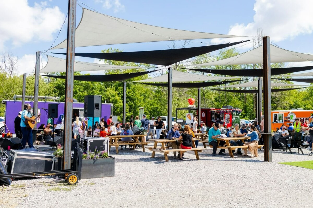 3 New Louisville Restaurants to Try (Plus 3 New Menus!) - 3 People sit at picnic tables and eat near food trucks in an outdoor area with sunshades, surrounded by greenery on a sunny day.