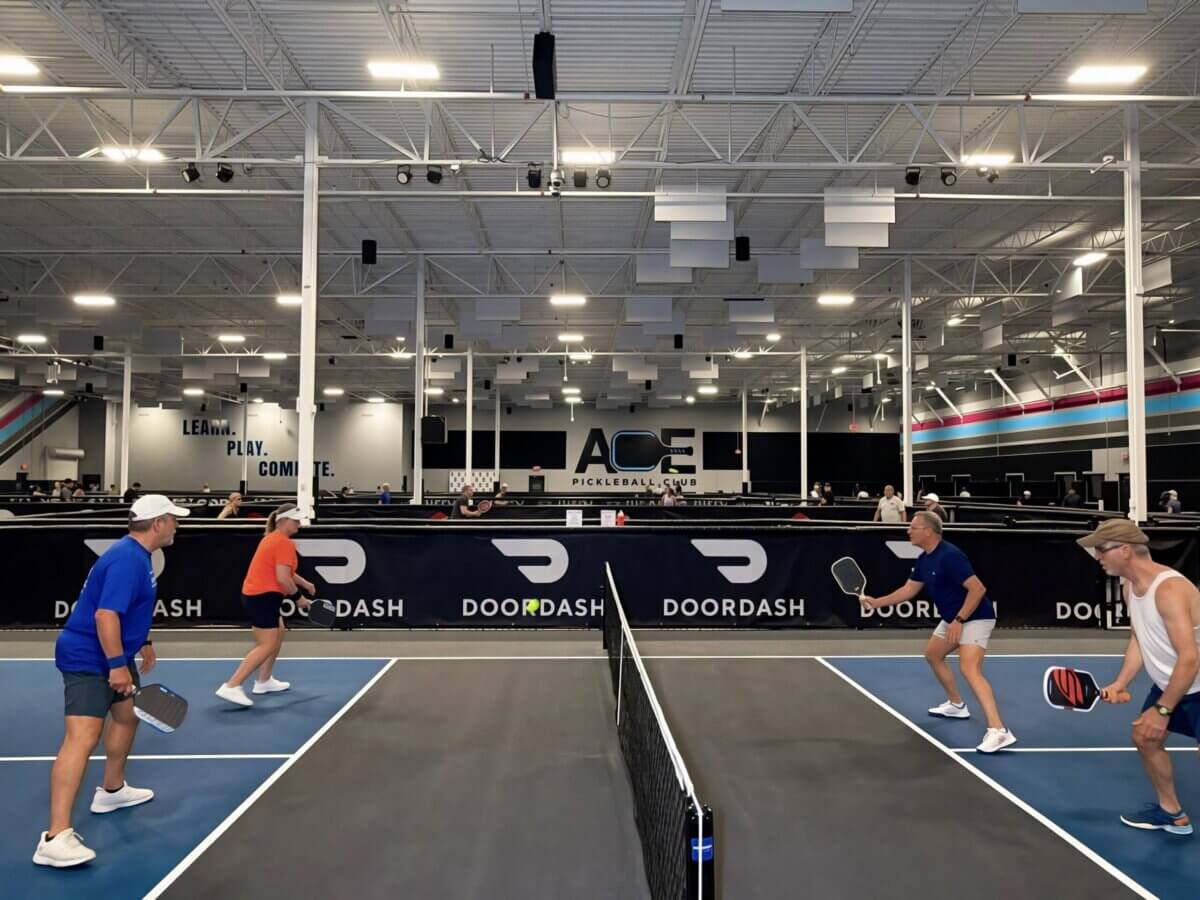 Four people play doubles pickleball on an indoor court in Louisville, with two on each side of the net under bright ceiling lights. A large “ACE” sign is visible in the background, highlighting one of the best places to play Pickleball in the city.