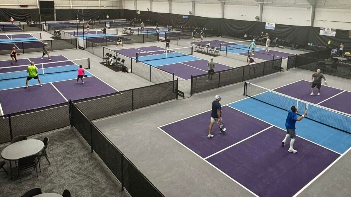 Players compete in games across multiple indoor pickleball courts, with tables and chairs seen in the foreground, making it one of the top places to play pickleball in Louisville.