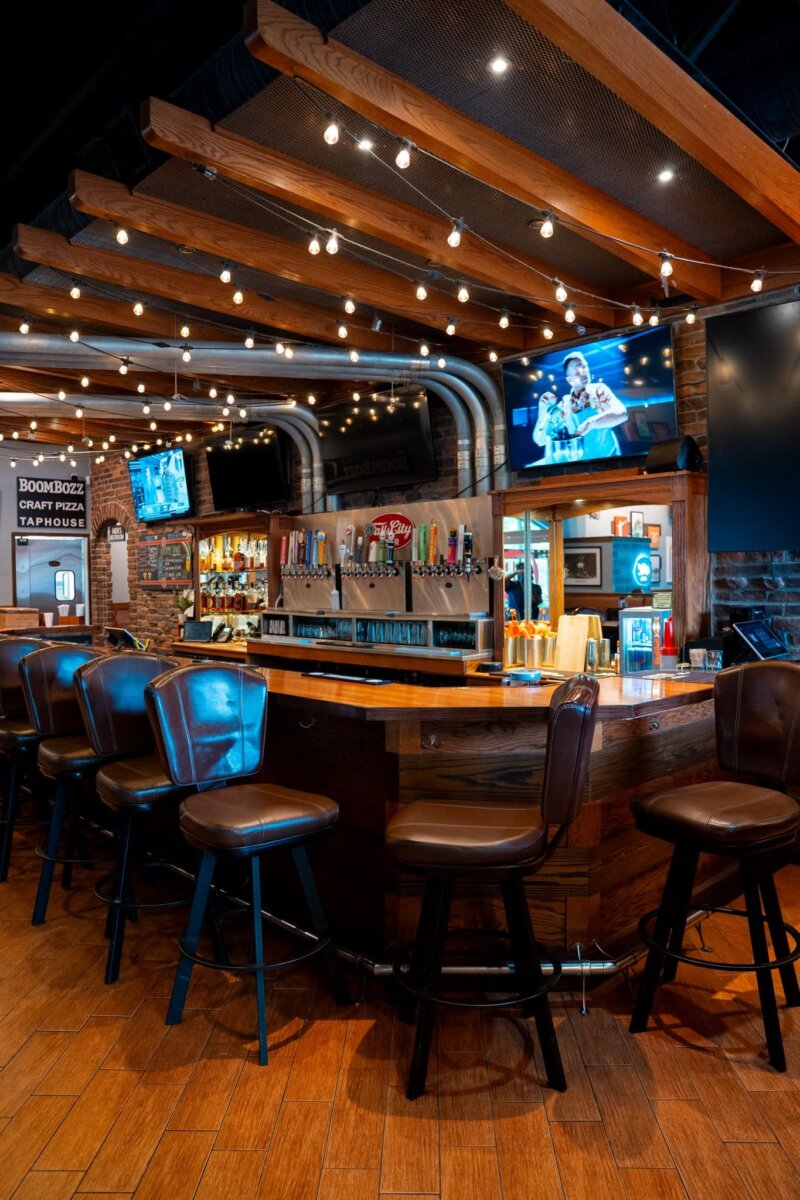 The Best Louisville Bars & Breweries for Watching Football - 2 A lively bar with wooden stools, multiple beer taps featuring selections from popular Louisville breweries, overhead string lights, mounted TVs perfect for watching football, and exposed brick walls.