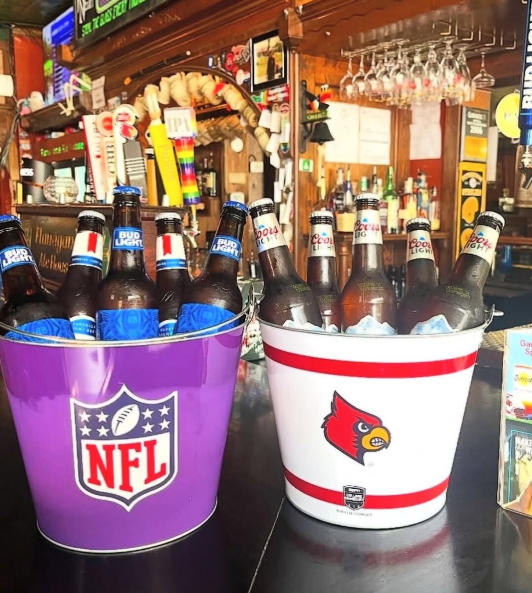 The Best Louisville Bars & Breweries for Watching Football - 3 Two buckets filled with bottled beer are on a bar counter, perfect for watching football with friends. One bucket has the NFL logo and the other features the Louisville Cardinals logo, celebrating local spirit and favorite teams. Draft beer taps and bar decor—reminiscent of popular Louisville bars—are visible in the background, creating a lively atmosphere ideal for game day gatherings.