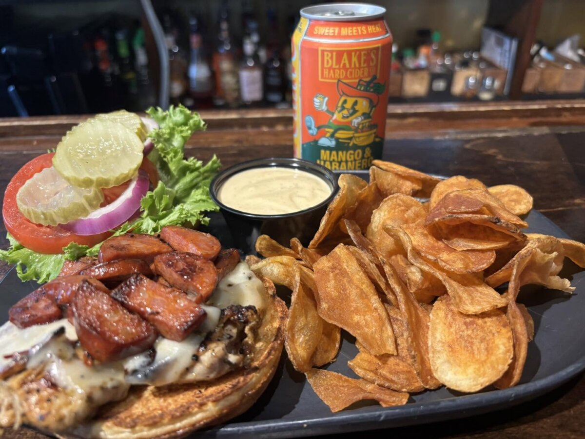 The Best Louisville Bars & Breweries for Watching Football - 4 Grilled chicken sandwich with cheese, sausage, lettuce, tomato, onions, pickles, house chips, dipping sauce, and a can of Blake’s Mango & Habanero cider on a black tray—perfect for watching football or enjoying at one of your favorite Louisville bars.
