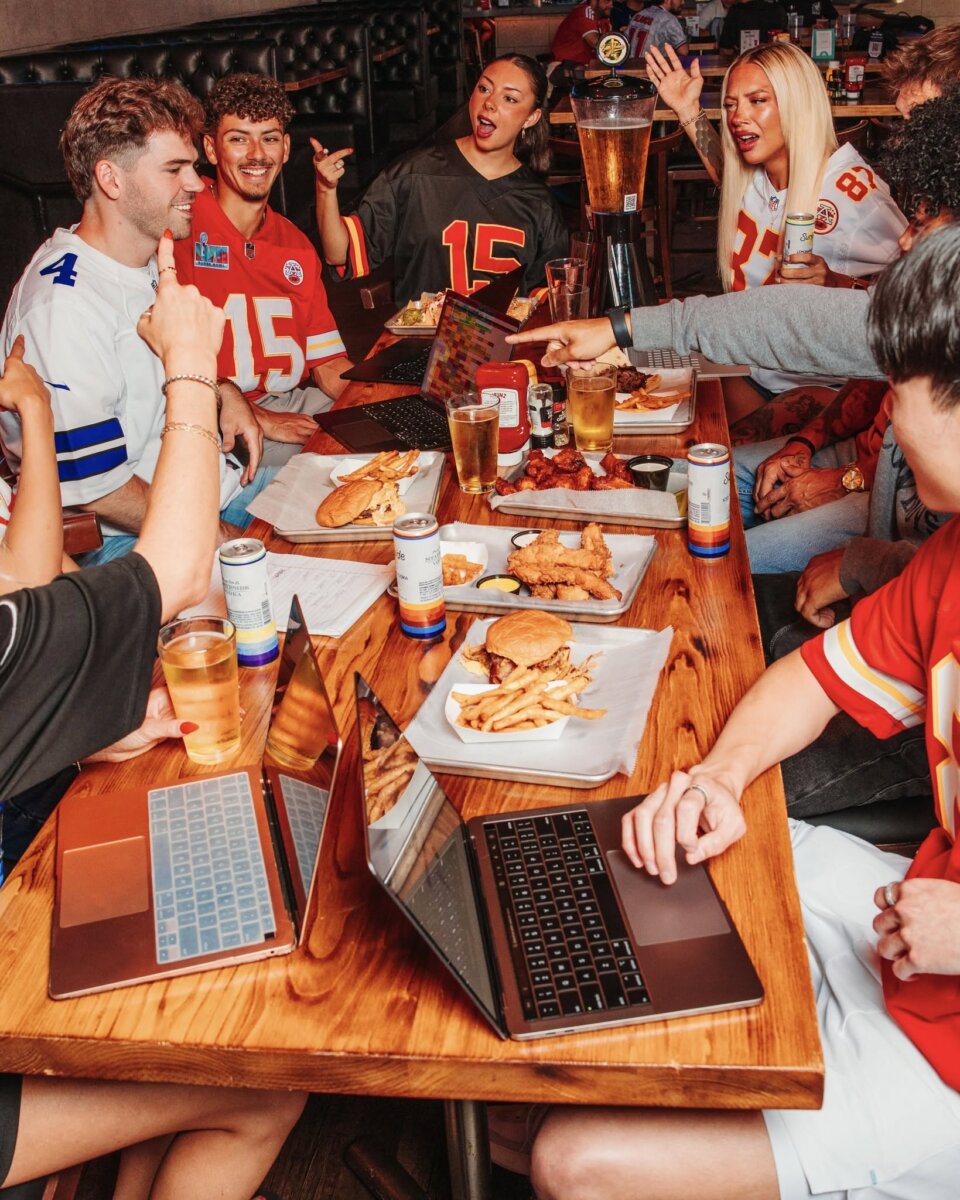 The Best Louisville Bars & Breweries for Watching Football - 5 Group of people wearing football jerseys sit around a table with food, drinks from local Louisville breweries, and laptops, watching football together and cheering.