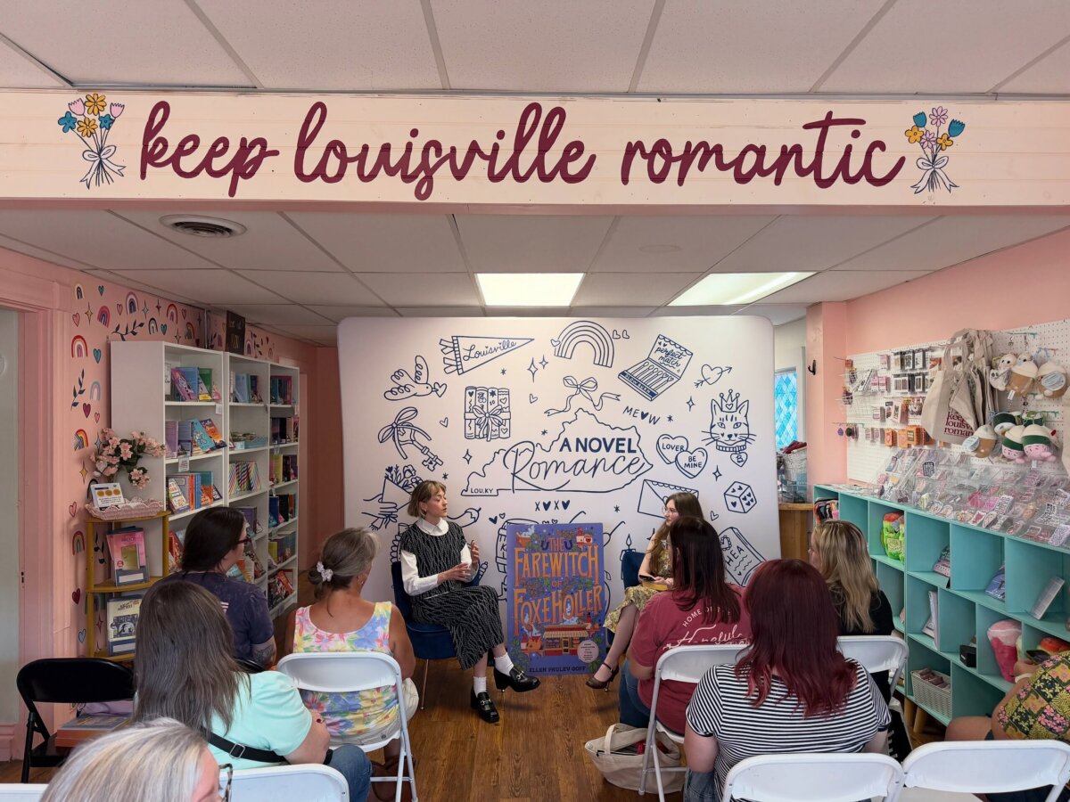 A small group listens to two speakers in a bookshop decorated with pastel colors, bookshelves, and a sign that reads “keep louisville romantic” above the stage area.