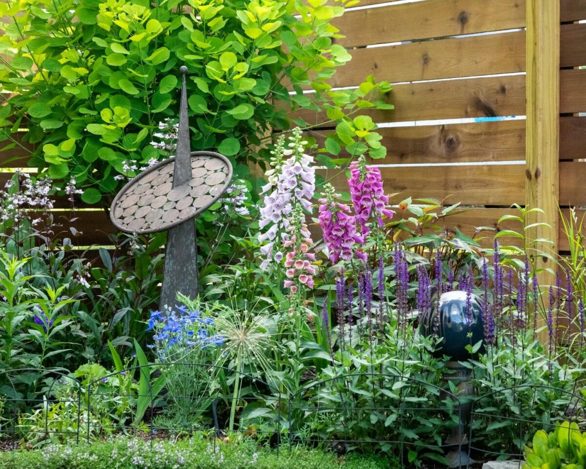 A garden in Louisville with various blooming flowers, a wooden fence, a round garden sculpture on a pole, and vibrant green foliage in the background—a perfect spot to relax and meet new people.