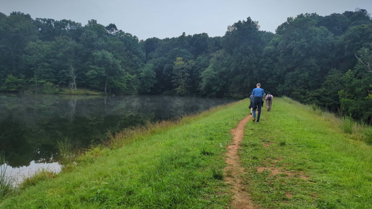 Three people walk along a narrow dirt path beside a calm lake in Louisville, bordered by green grass and dense trees under an overcast sky.
