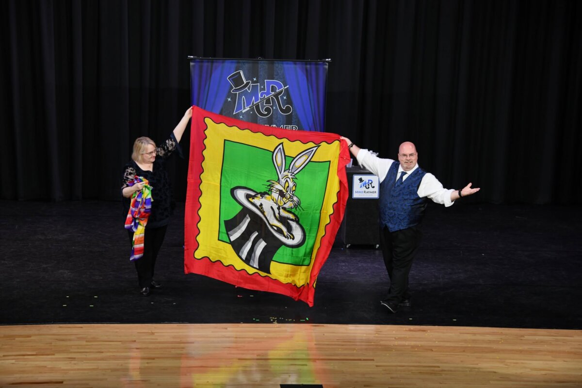 Two performers stand on stage in Louisville holding a large colorful scarf with a cartoon rabbit in a hat, in front of a banner that reads “M&C.” The lively scene sets the perfect backdrop for social activities and opportunities to meet new people.