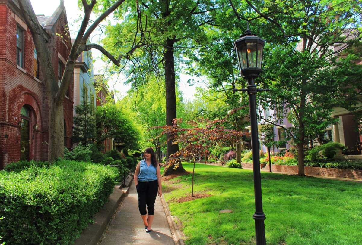 A woman walks along a shaded sidewalk in a residential Louisville neighborhood with brick buildings, lush greenery, and a lamppost, enjoying the welcoming atmosphere and opportunities to meet new people.