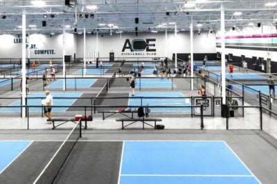 Indoor pickleball facility in Louisville with multiple blue courts, where players are actively competing and “ACE Pickleball Club” signage stands out on the wall. Bright overhead lighting illuminates one of the top places to play pickleball in the city.