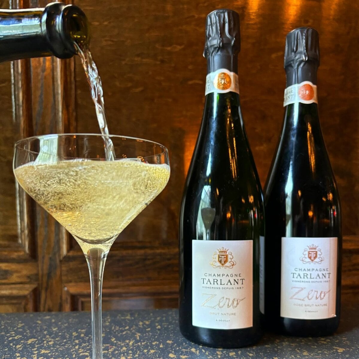 8 Louisville Wine Bars We Love - 2 Champagne is poured into a glass next to two bottles of Tarlant Zero Brut Nature champagne on a counter with a wooden background.