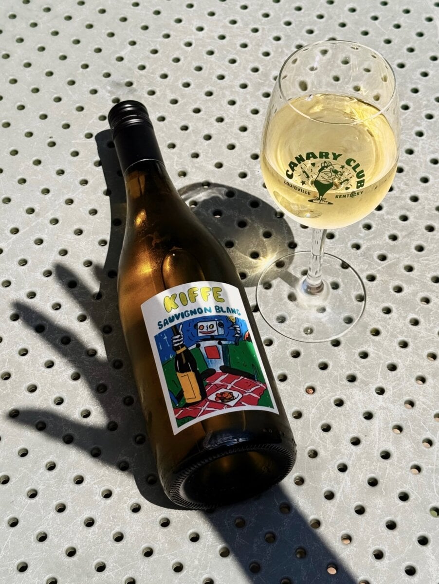 8 Louisville Wine Bars We Love - 3 A bottle of Kiffe Sauvignon Blanc and a glass of white wine sit on a perforated metal table in sunlight.
