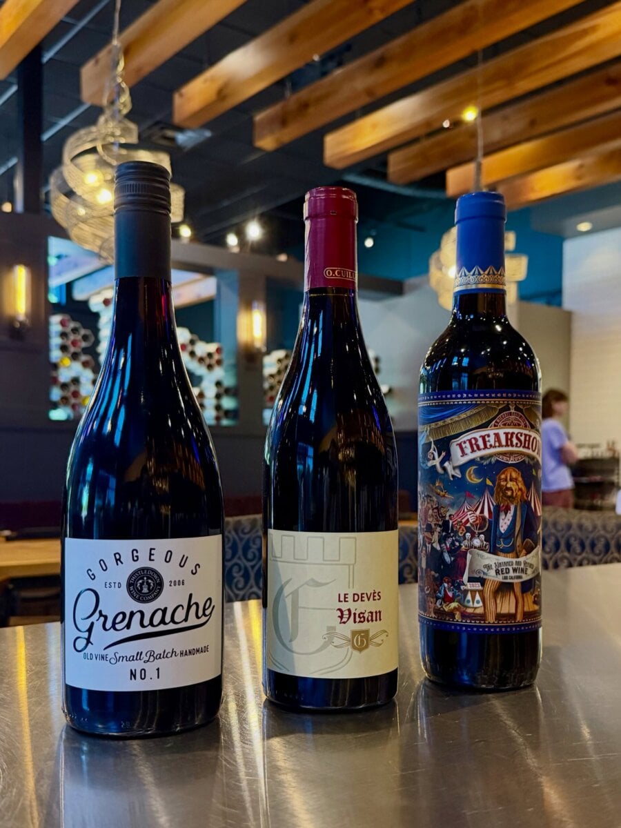 8 Louisville Wine Bars We Love - 4 Three bottles of red wine are displayed on a metal table in a modern restaurant with wooden ceiling beams and wine racks in the background.