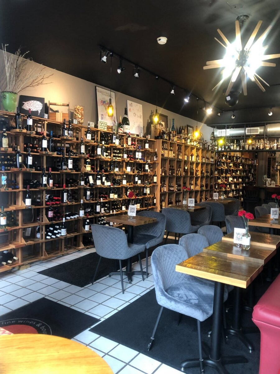 8 Louisville Wine Bars We Love - 5 Experience the inviting ambiance of a wine shop interior that rivals the best wine bars Louisville has to offer, with shelves full of wine bottles, small tables with gray chairs, white tile flooring, and modern ceiling lights—perfect for enjoying Louisville nightlife in style.