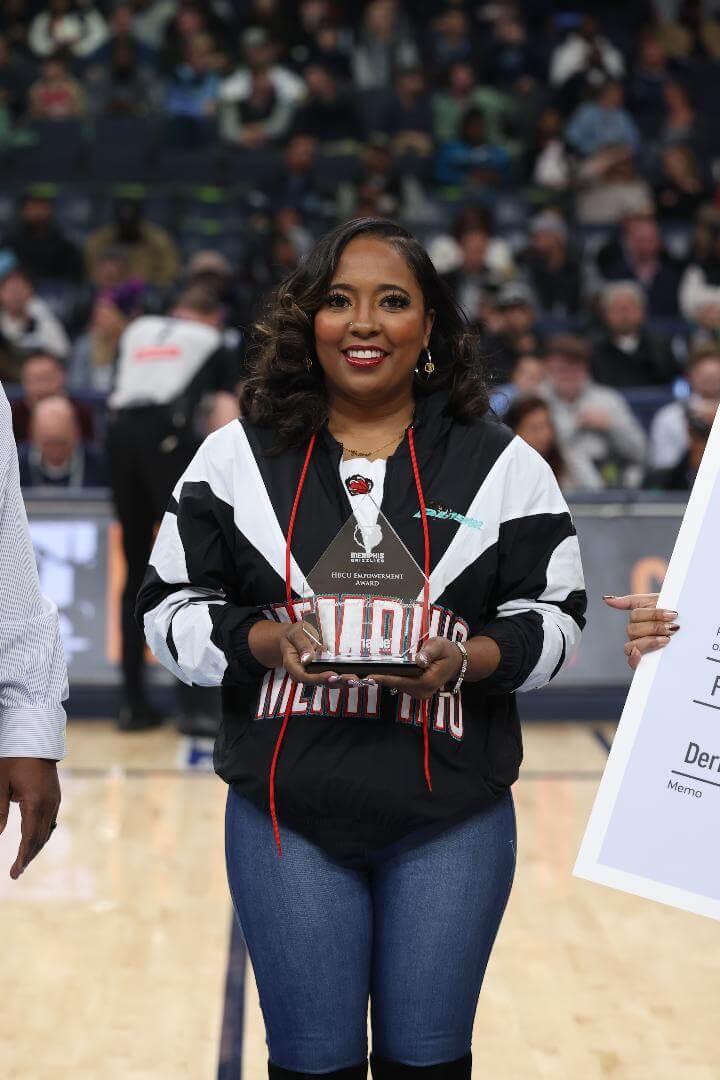 A woman in a Memphis jacket holds an award plaque while standing on a basketball court, with a crowd seated in the background.