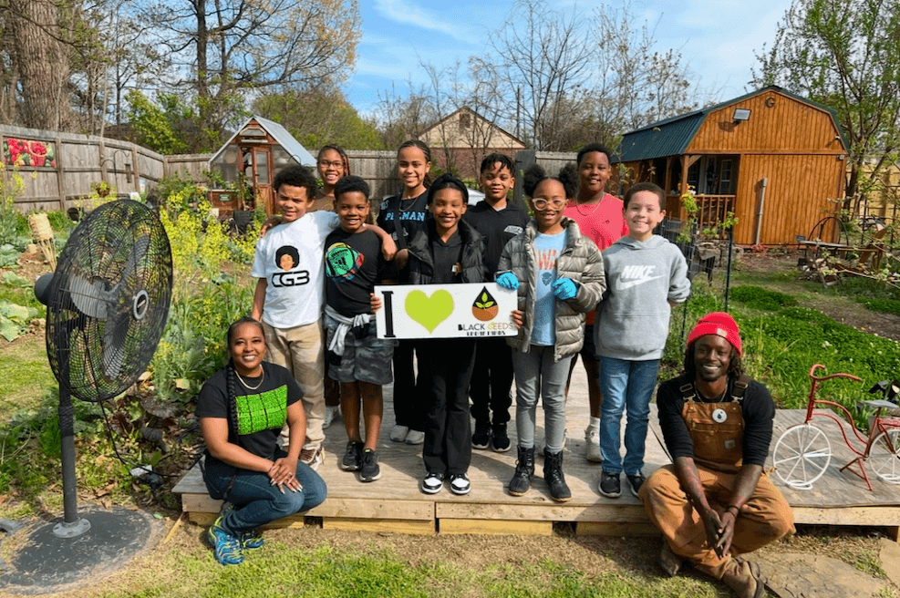 A group of children and two adults pose outdoors on a wooden platform in a garden, holding a sign that says “I ♥ Black Kids Do Garden.” Among them is Derravia Rich.