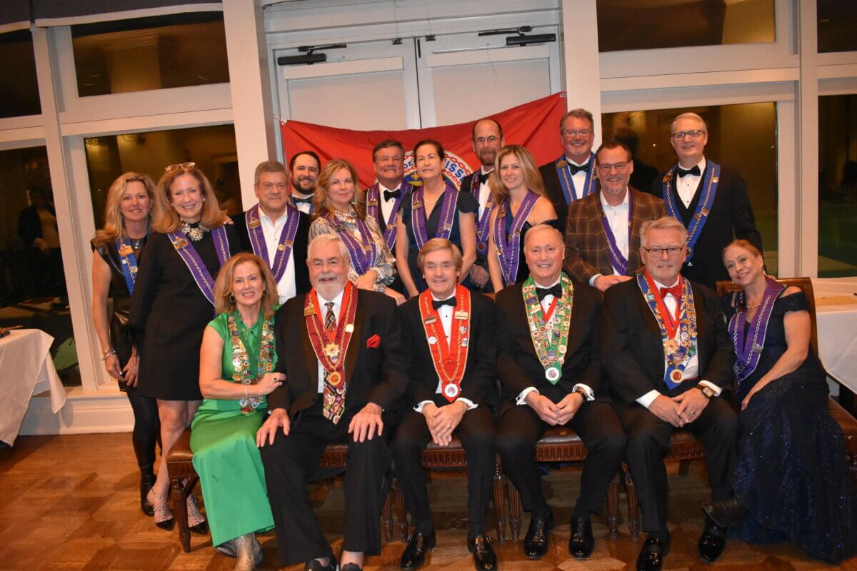 A group of men and women in formal attire pose indoors, some seated and some standing, wearing ceremonial sashes and medallions in front of a red flag. Among them is Pamela Hauber.