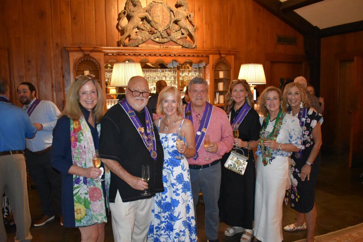 A group of seven adults, including Pamela Hauber, dressed in semi-formal attire, pose together holding drinks at an indoor social event with wood-paneled walls.