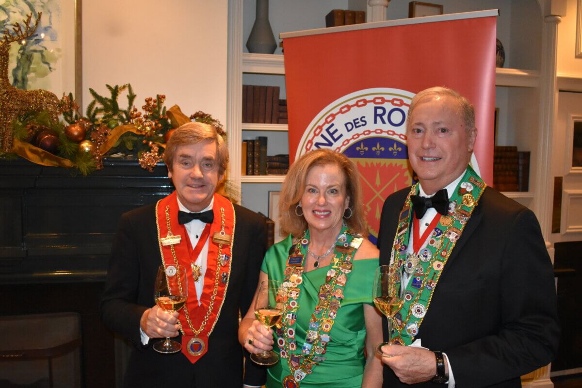 Three elegantly dressed adults, including Pamela Hauber, wearing ceremonial sashes and medals, pose indoors holding glasses of white wine.